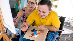 An older woman assists a man with down syndrome with a painting.